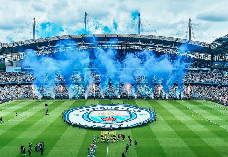 Manchester City Football stadium filled with blue smoke effects and fans surrounding the pitch.